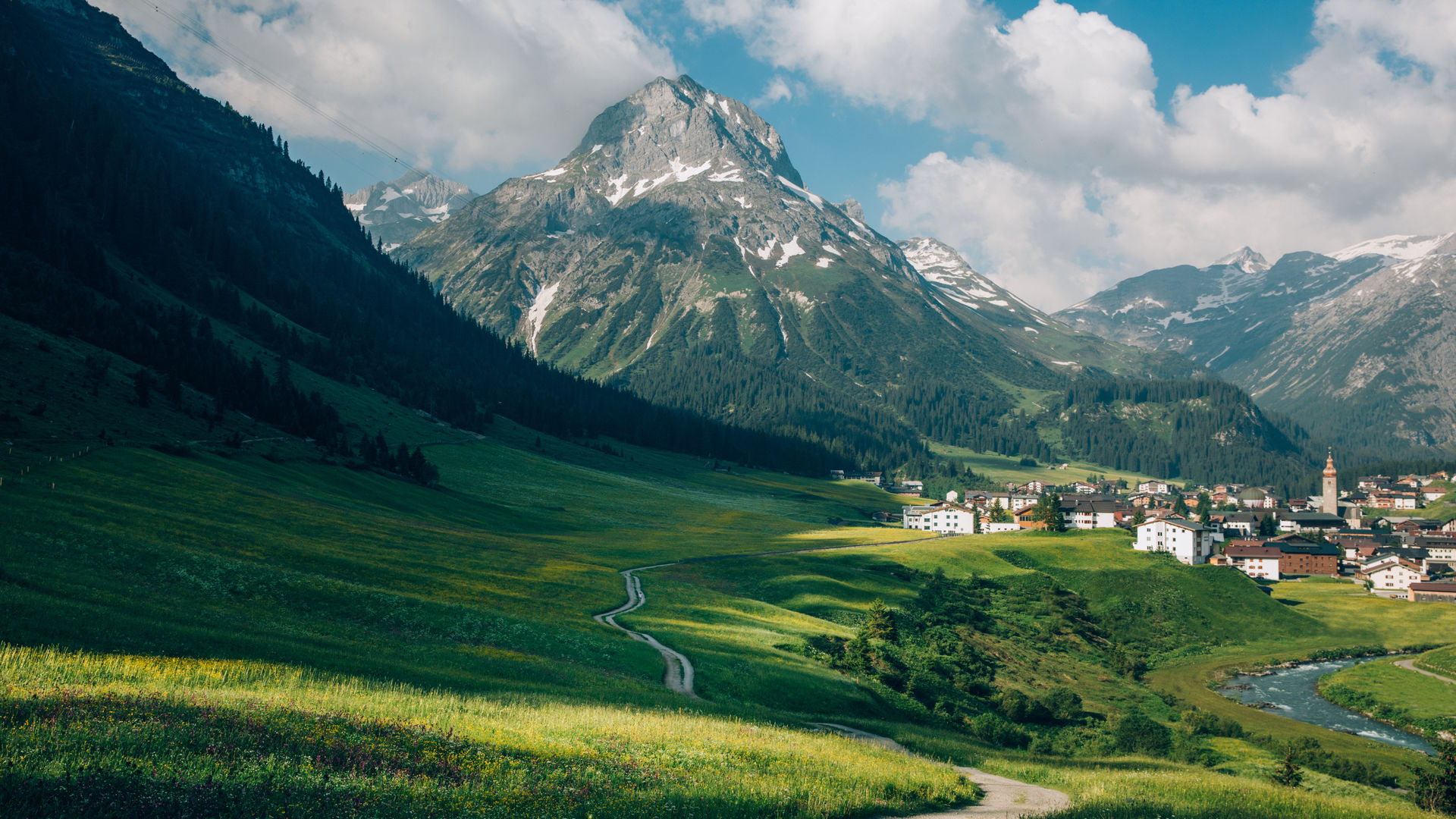 Wanderweg durch eine blühende Frühlingswiese, im Hintergrund sieht man das Dorf und den Fluss Lech und ein atemberaubendes Bergpanorama