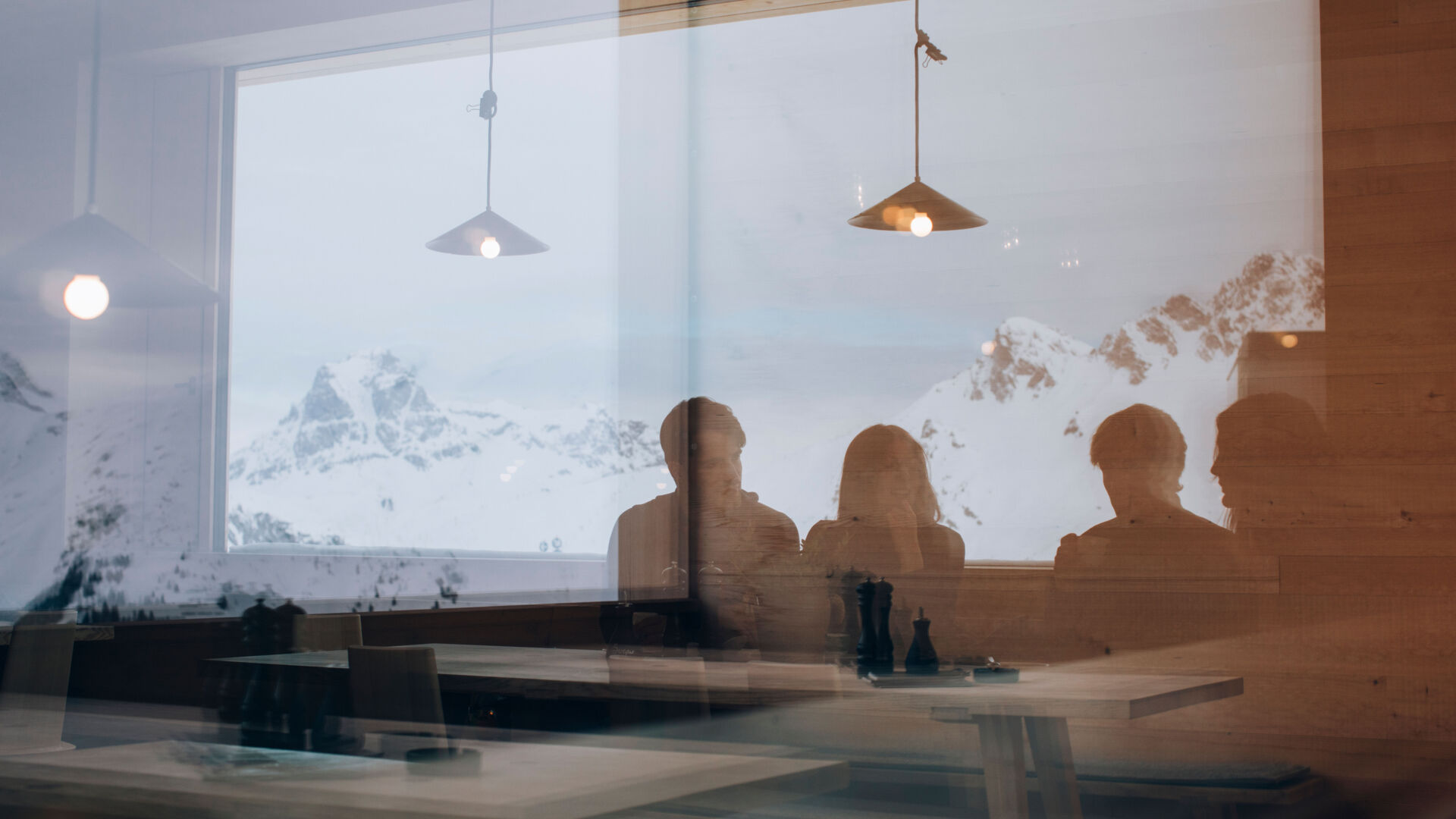 Blick in die Gaststube wo mehrere Personen eine gemütliche Zeit miteinander verbringen während die verschneiten Berge sich im Fenster widerspiegeln