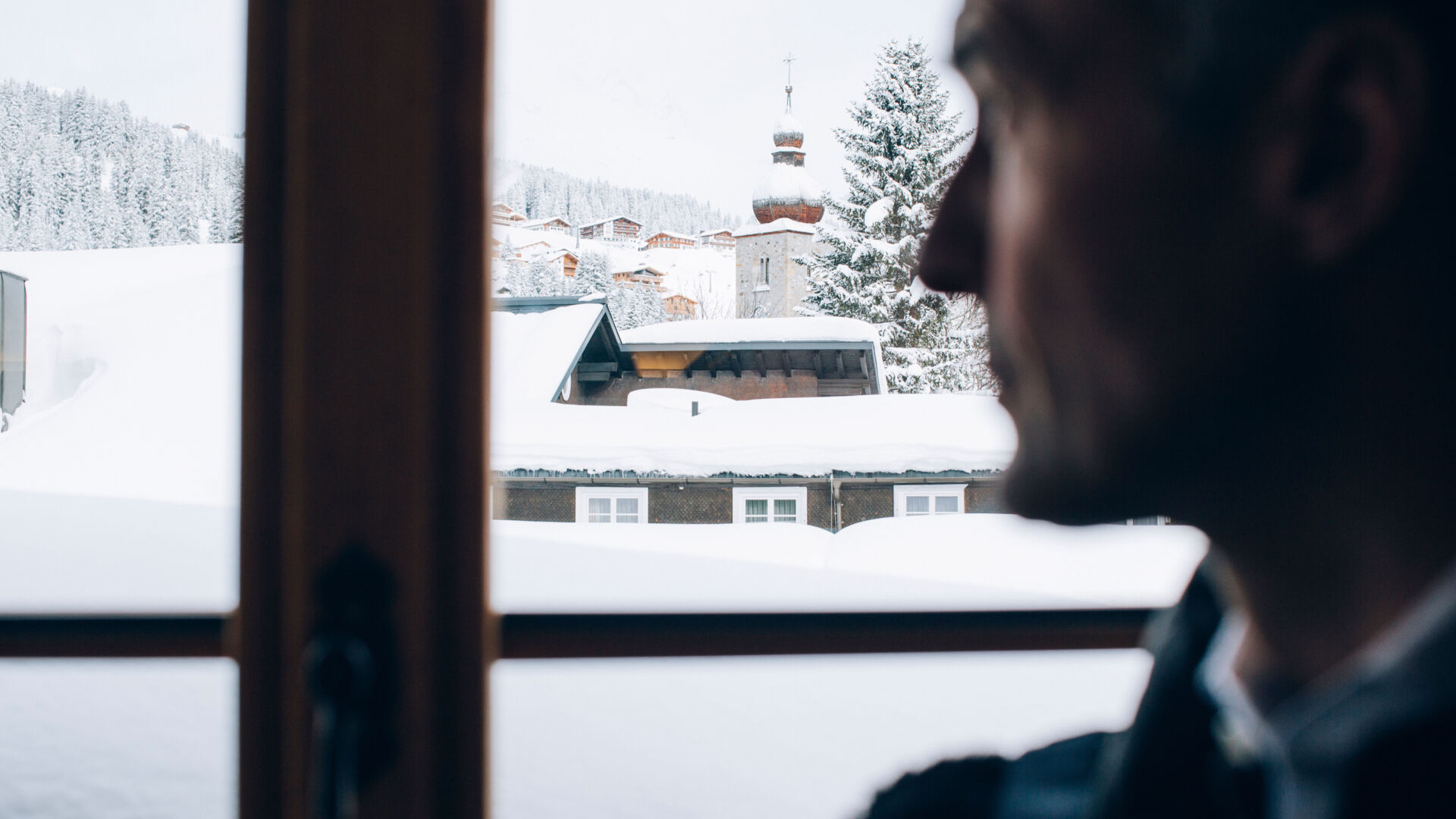 ein Mann blickt aus dem Fenster auf die schneebedeckten Dächer von Lech, im Hintergrund sieht man die winterlichen Wälder am Barg