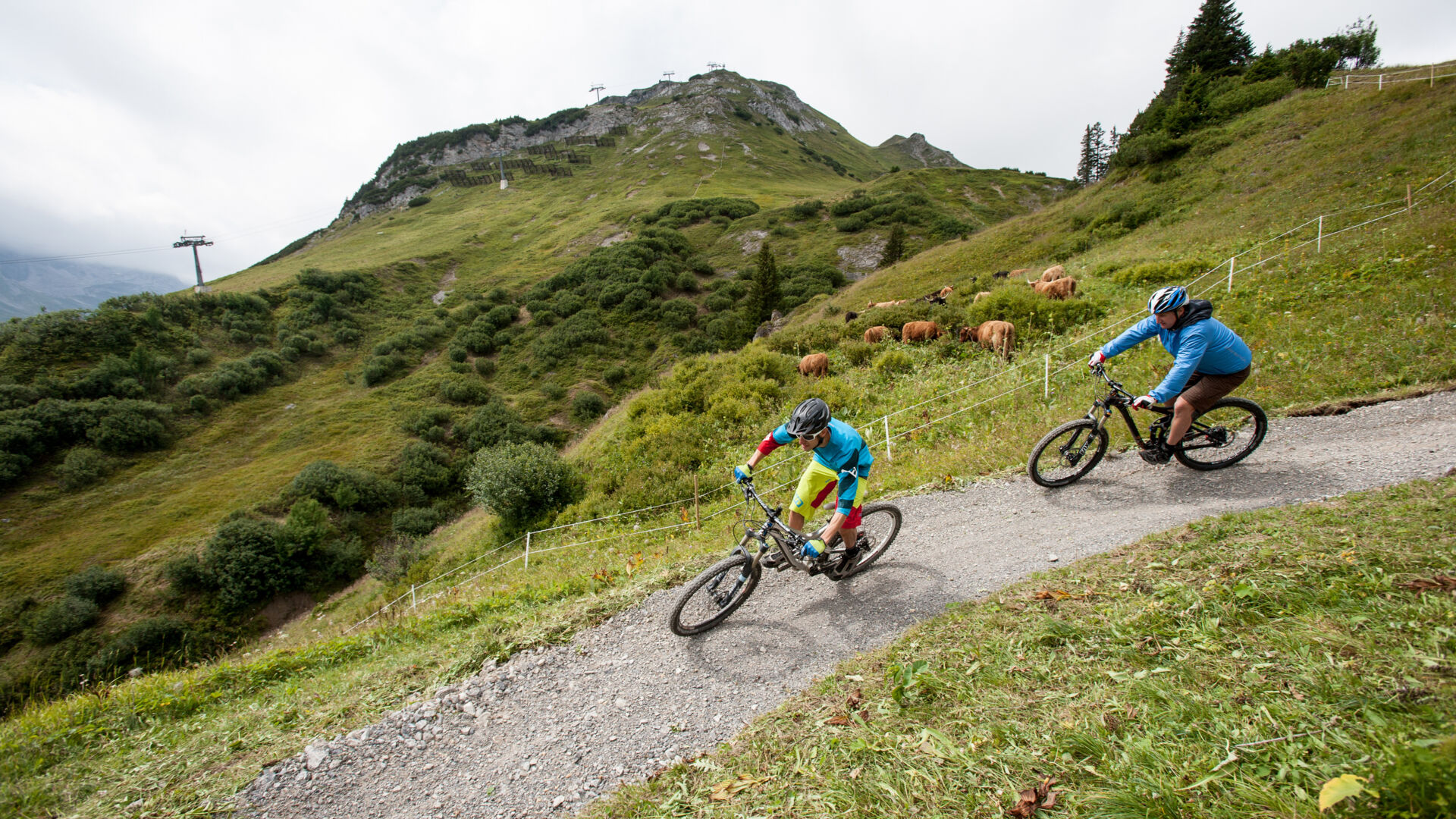 Zwei Radfahrer fahren einen Schotterweg im Gebirge hinab. Im Hintergrund erstrecken sich die weitläufigen Berge. 