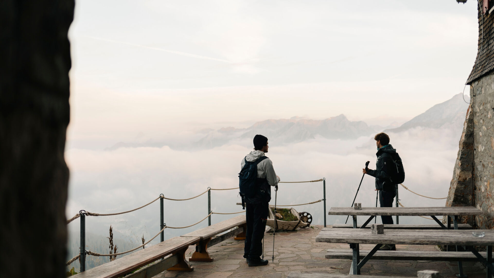 Zwei Wanderer stehen auf der Terrasse einer Hütte und blicken auf das weitläufige Bergpanorama und die tieferliegenden Wolken.