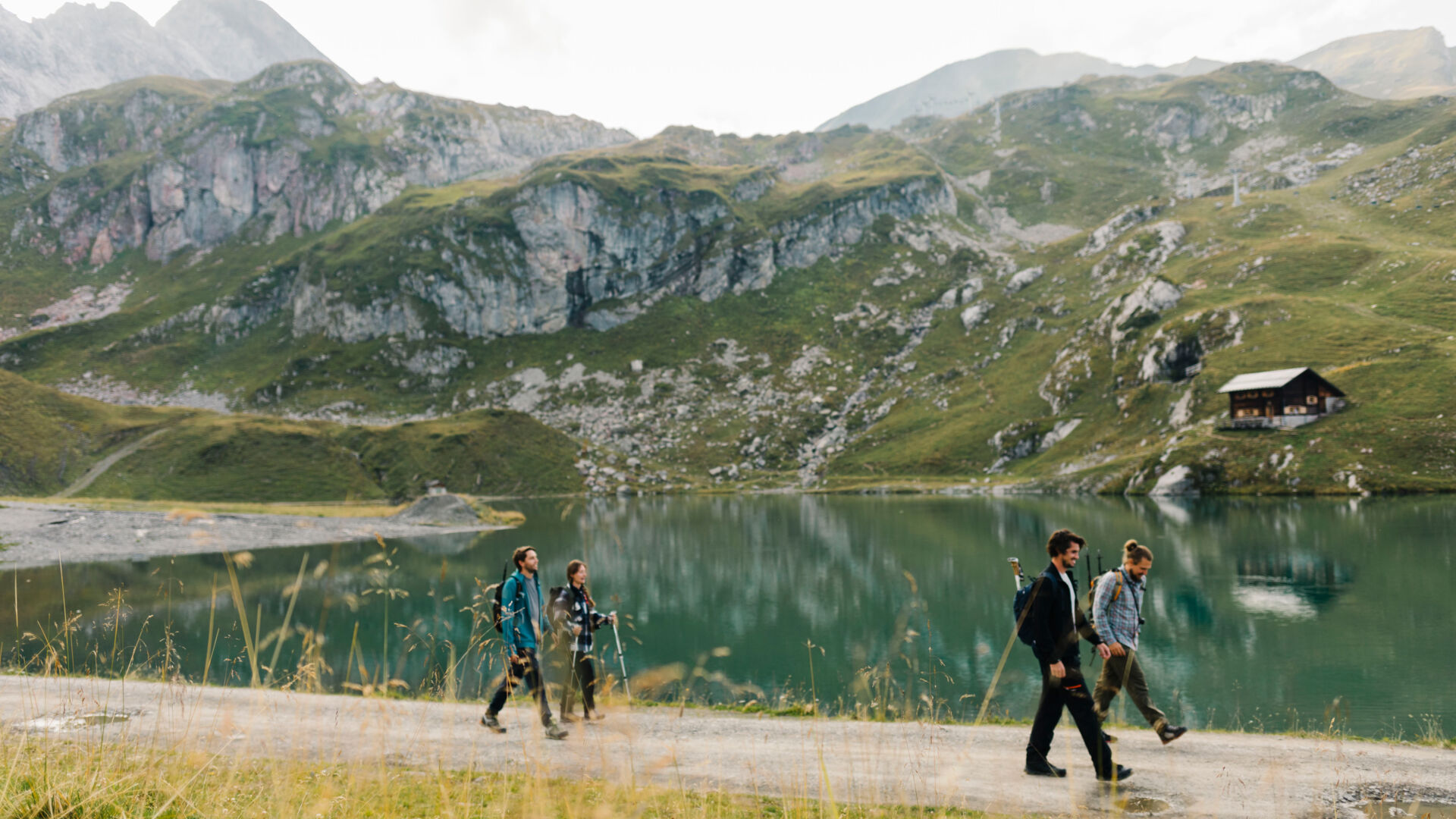 Wanderer gehen entlang eines Bergsees. Im Hintergrund sieht man ein weitläufiges Gebirgspanorama mit einer kleinen Hütte.
