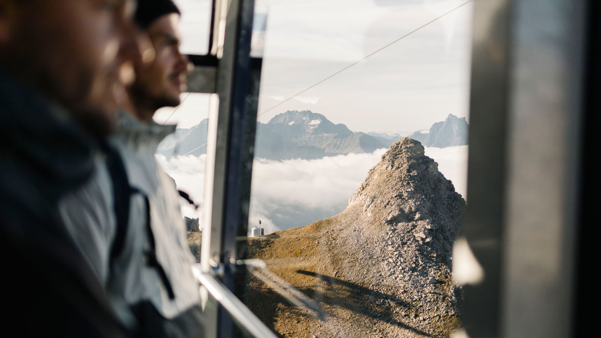 Zwei Wanderer blicken aus der Gondel hinaus auf die umliegenden Berge