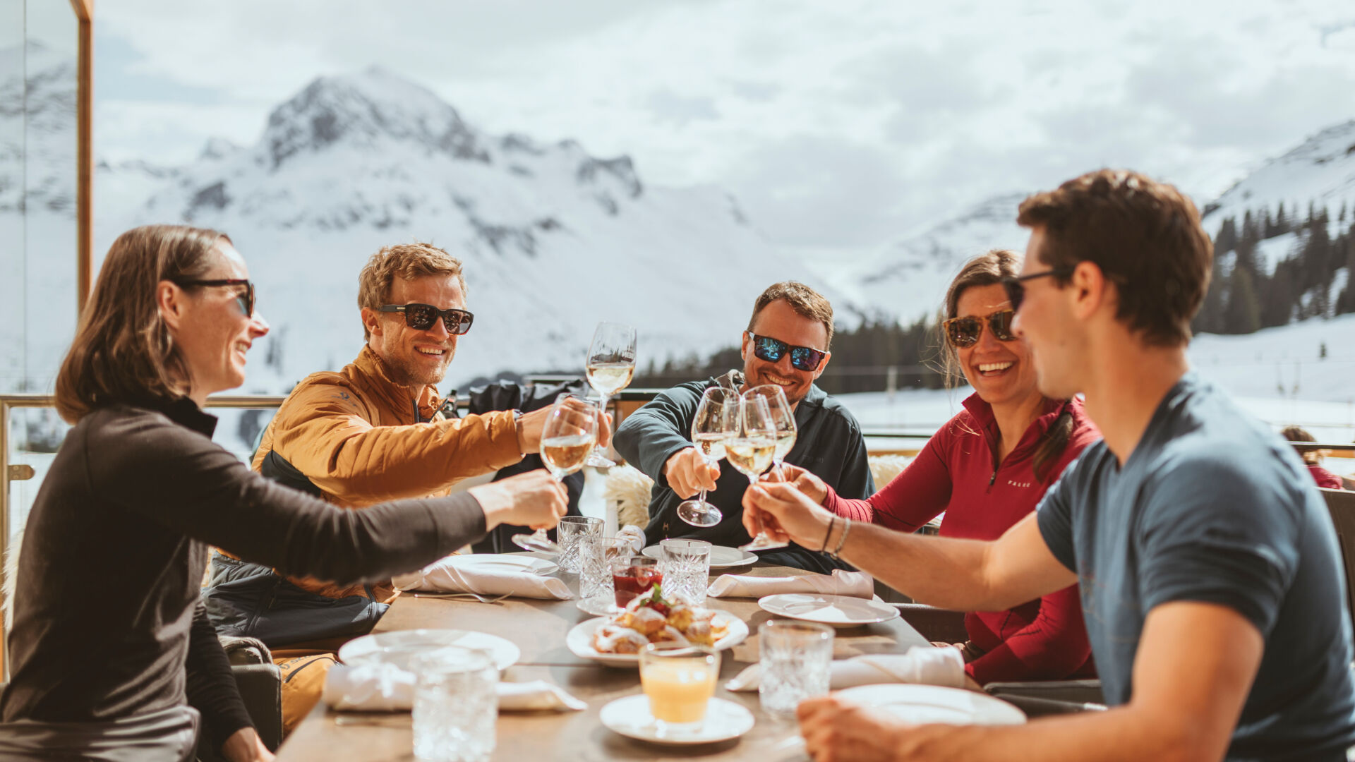 einige Freunde stoßen gemeinsam auf der Sonnenterrasse miteinander an, während sie sich kulinarisch verwöhnen und ein traumhaftes Bergpanorama im Hintergrund zu sehen ist