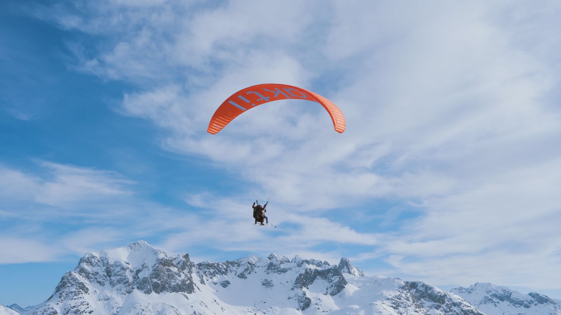 Tandemflieger fliegt vor blauem leicht bewölktem Himmel und im Hintergrund ist eine schneebedeckte weiße Bergkulisse zu sehen