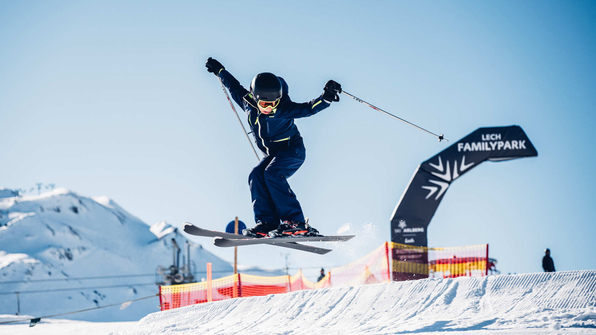 Junger Skifahrer springt mit gekreuzten Ski über einen Kicker im Lech Familypark, im Hintergrund Berge und blauer Himmel.