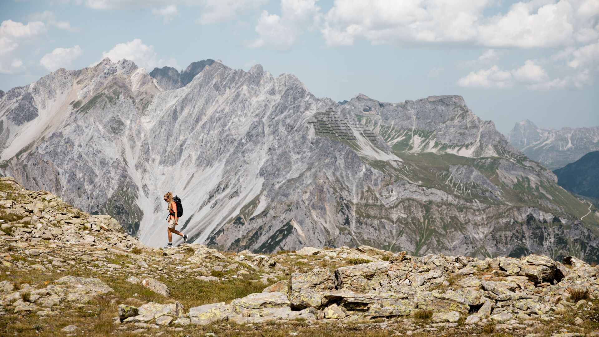 Wanderin mit Rucksack geht über einen felsigen Bergrücken, im Hintergrund schroffe Gipfel der Lechtaler Alpen.