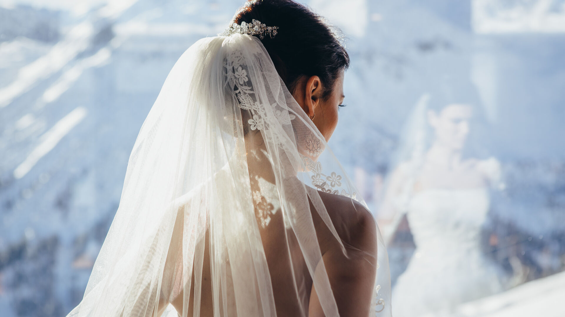 Man sieht die Braut von hinten, wie sie aus dem Fenster auf die verschneite Bergkulisse im Winter blickt