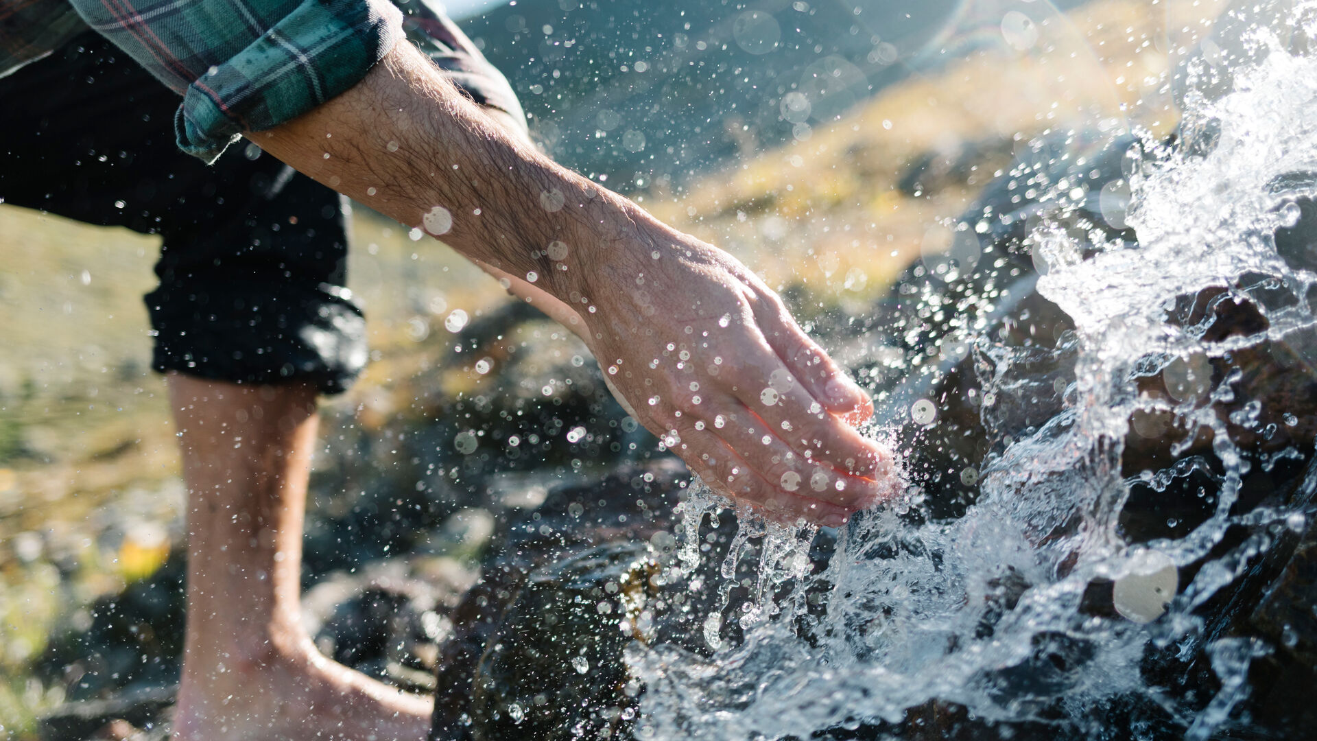 Ein Mann steht barfuß in einem Bach und berührt mit den Händen das klare Bergwasser