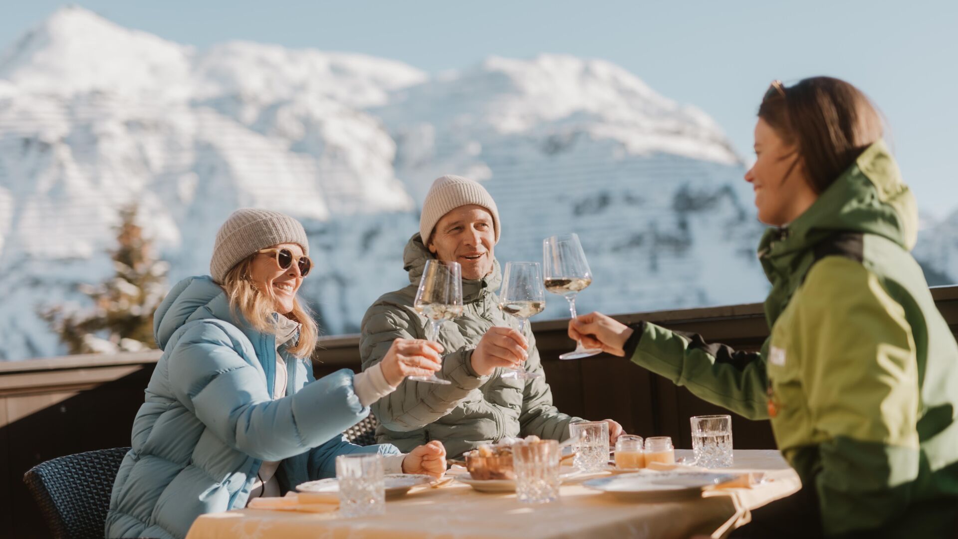 drei Freunde genießen auf der Sonnenterrasse die winterliche Berglandschaft und die Kulinarik von Lech Zürs mit einem Glas Wein