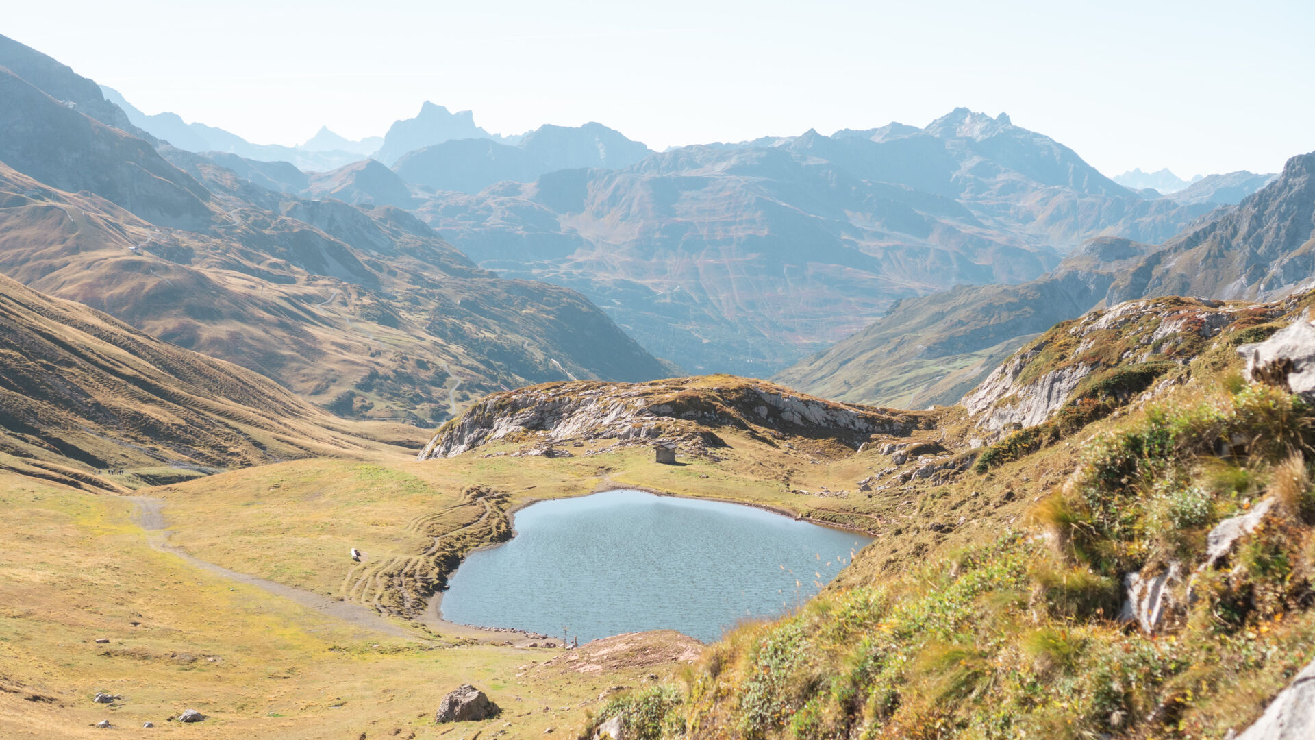 Blick auf einen kleinen Bergsee mit einer Steinhütte am Ufer, umgeben von herbstlich gefärbten Almwiesen und Felsen, im Hintergrund mehrere dunstige Bergketten.