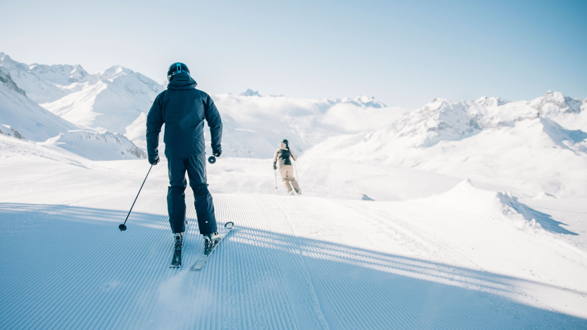 zwei Skifahrer fahren über die frischgewalzte Piste bei Sonnenschein hinunter, im Hintergrund sieht man die traumhafte Bergkulisse im Winter