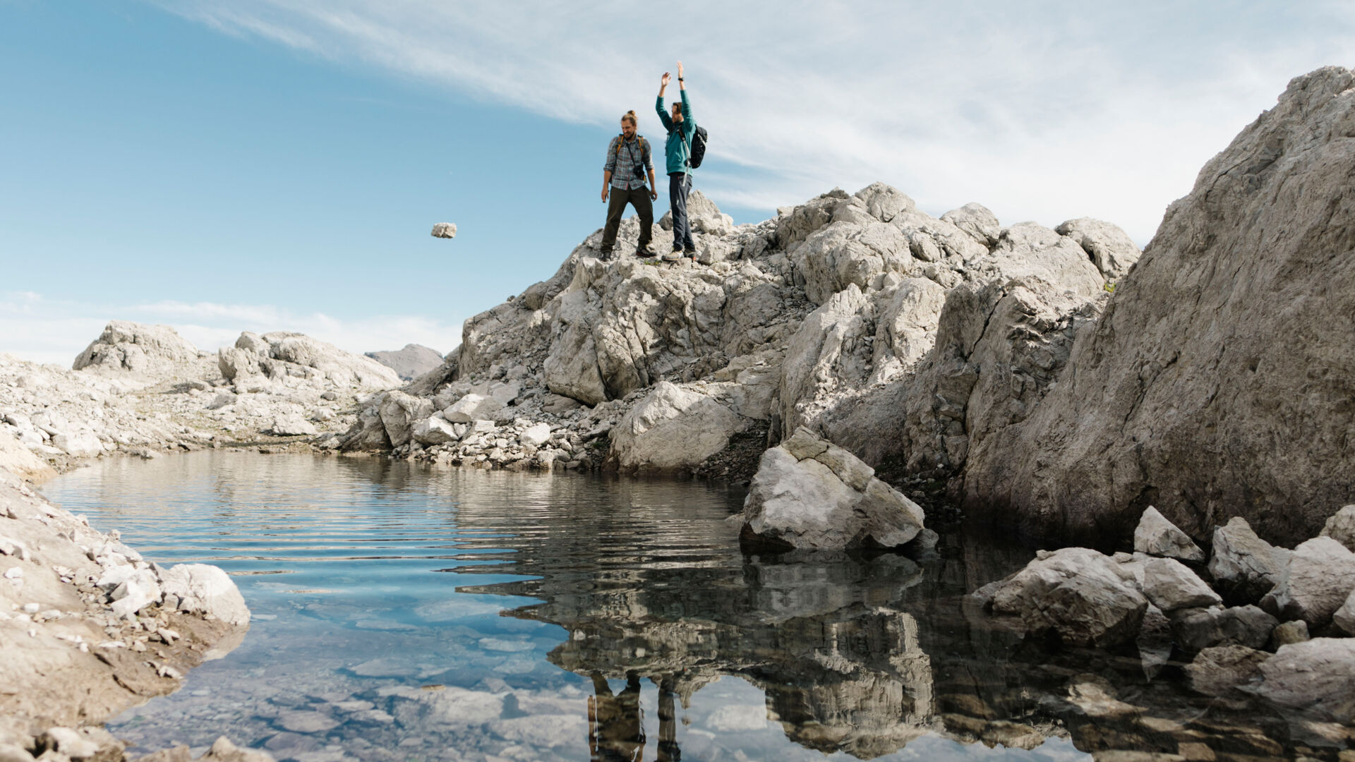 Zwei Wanderer stehen vor einen kleinem Gebirgssee und einer der beiden wirft einen Stein ins Wasser. 