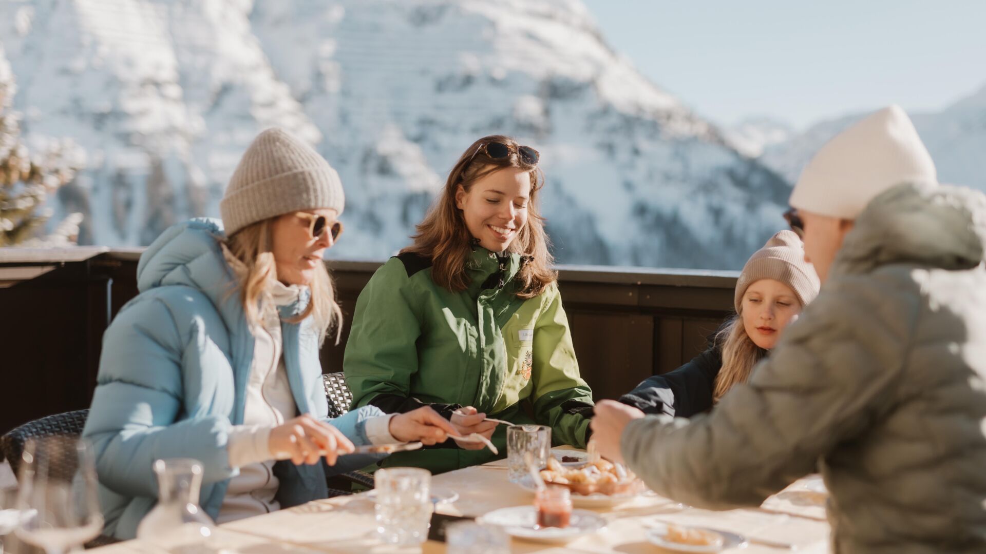 eine Familie genießt den warmen Kaiserschmarren auf der Sonnenterrasse mit einem atemberaubenden, verschneiten Bergpanorama in Lech Zürs am Arlberg