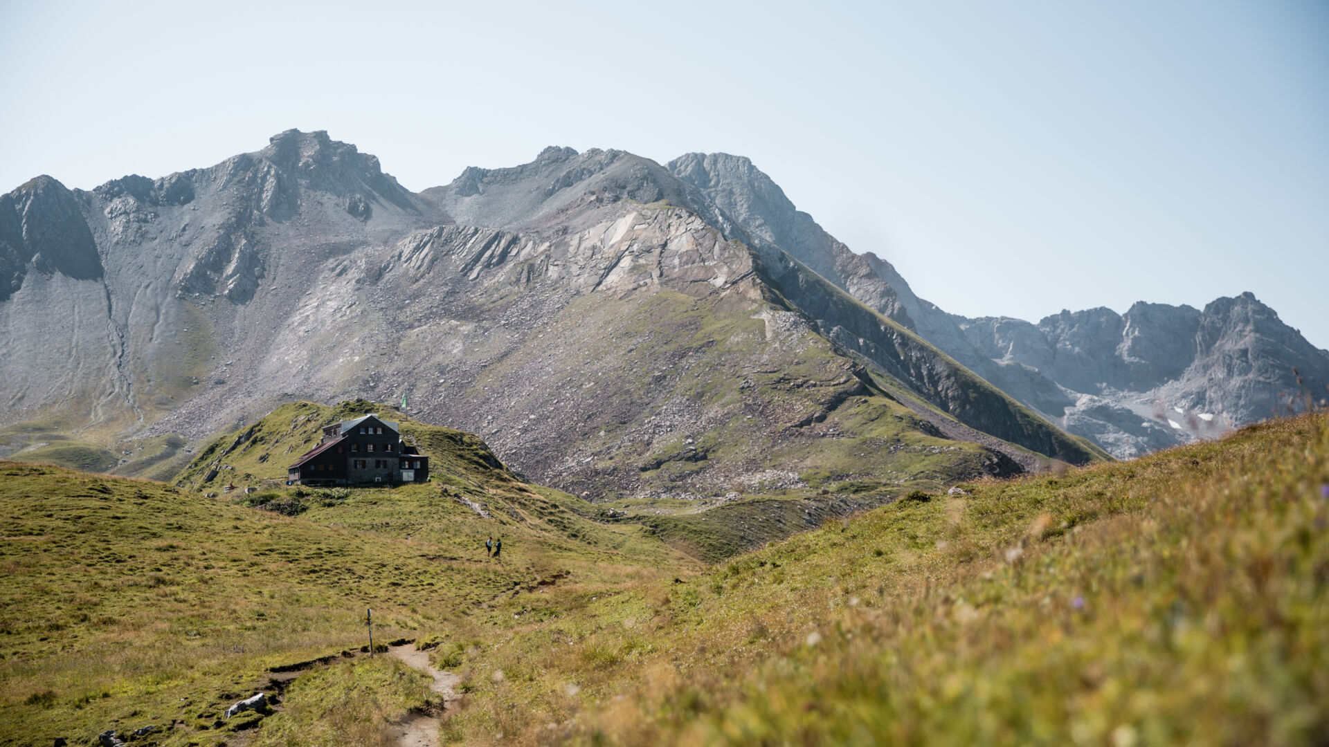 Die Stuttgarter Hütte liegt auf einer grünen Bergkuppe vor schroffem Felsmassiv, ein Wanderweg führt darauf zu.