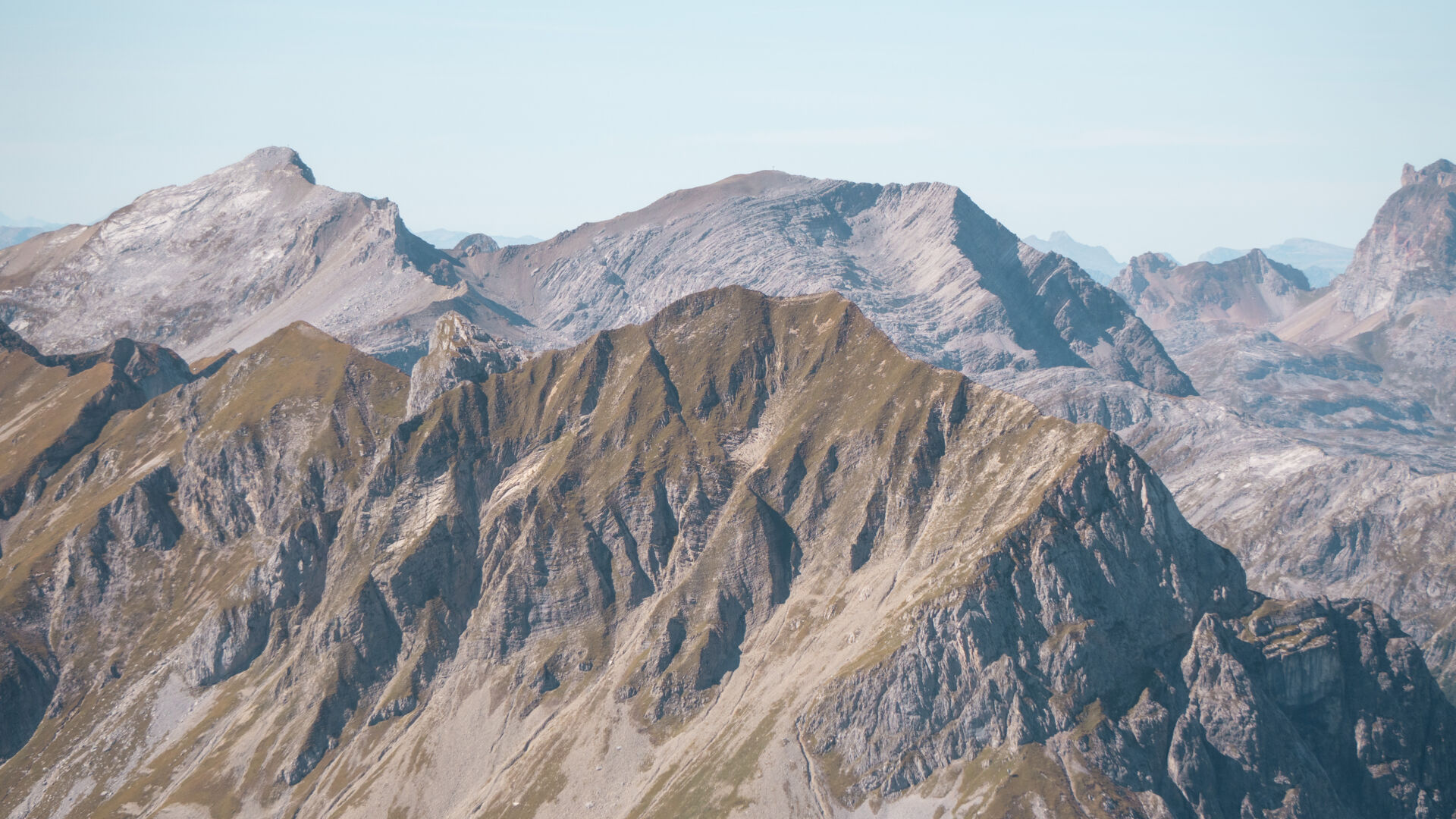 Zerklüftete Bergkette im Herbst mit felsigen Graten und herbstlich gelbbraunen Grashängen, dahinter hellere, graue Gipfel unter einem klaren, hellblauen Himmel.