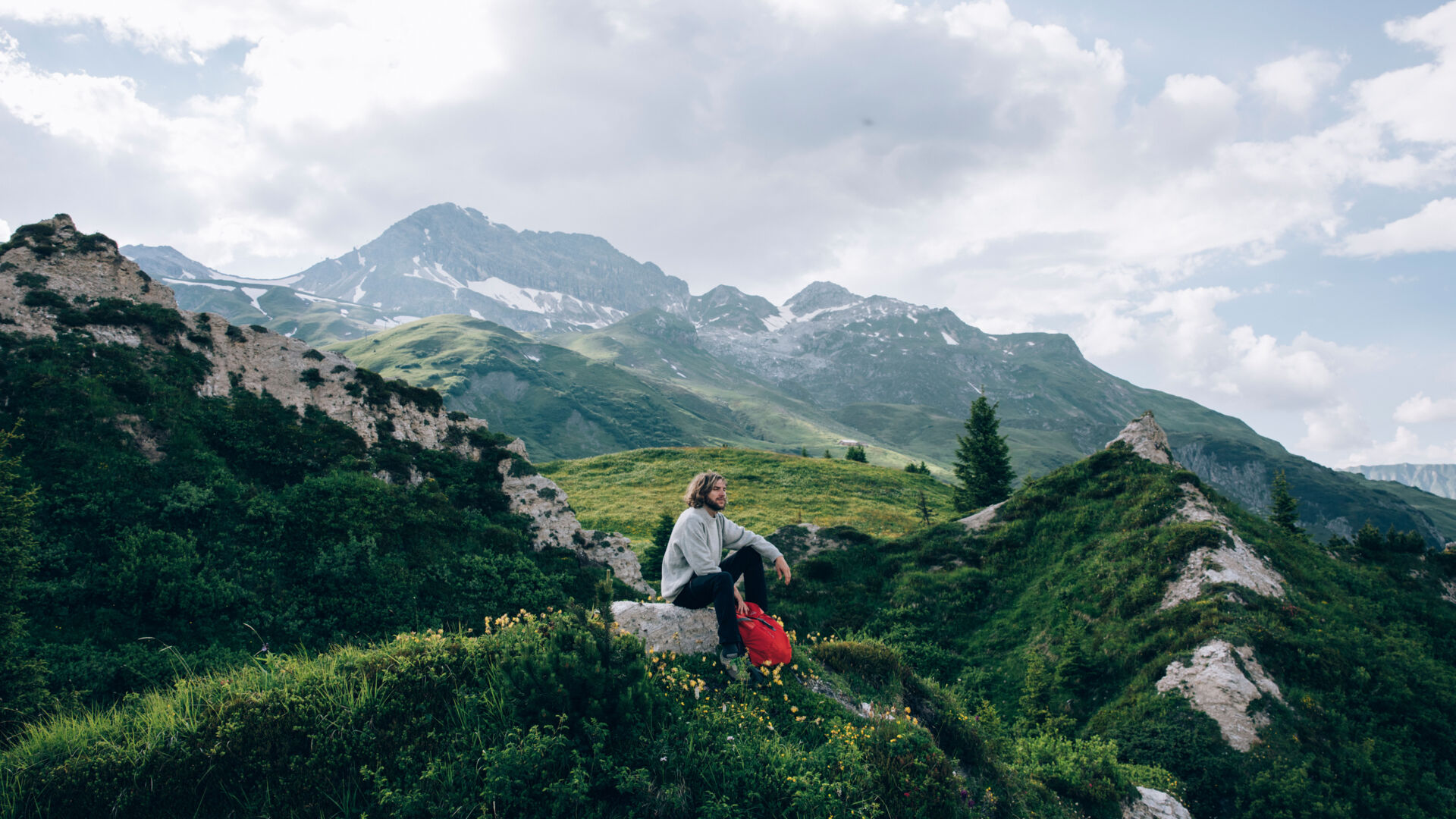 ein Wanderer macht eine Pause und genießt das traumhafte Bergpanorama inmitten von saftig grünen Wiesen