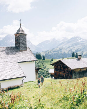 Blick auf die Kirche und urige Holzh&auml;user in B&uuml;rstegg, w&auml;hrend zwei Wanderer auf dem Wanderweg laufen, im Hintergrund ist das leicht bew&ouml;lkte Bergpanorama zu sehen