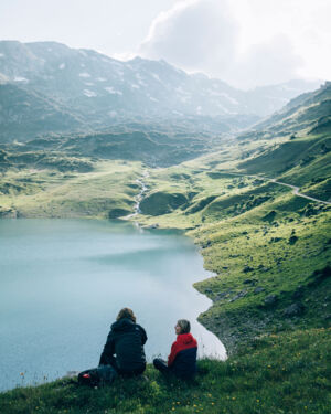 zwei Wanderer sitzen in der bl&uuml;henden Bergwiese und genie&szlig;en die Aussicht auf den Formarinsee, im Hintergrund erstrecken sich die felsigen Berggipfel