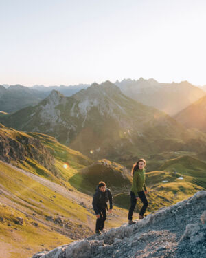 Eine Frau und ein Mann wandern bergauf während im Hintergrund die Sonne über die Berggipfel scheint