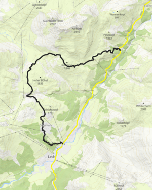 zwei Wanderer erreichen über eine Forststraße Bürstegg, neben den blühenden Bergwiesen sieht man im Hintergrund die steinigen Bergwiesen