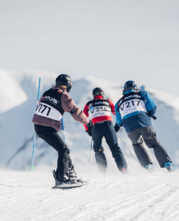 Zwei Skifahrer und ein Snowboarder fahren mit ihren Startnummern-Bibs bei strahlend blauem Himmel die verschneite Strecke des Weißen Rings hinunter