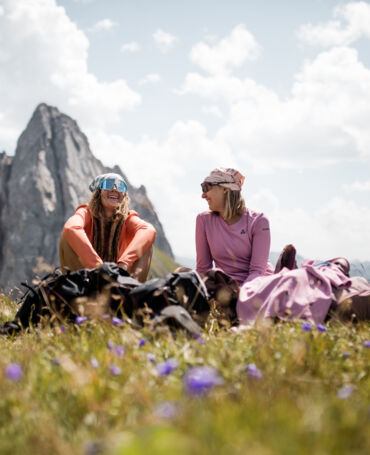 Zwei lachende Wanderinnen sitzen zwischen Rucksäcken auf einer blühenden Bergwiese, im Hintergrund ein spitzer Felsgipfel.