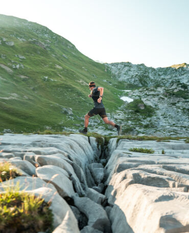 Trailrunner springt über eine tiefe Felsspalte im Karstgestein, im Hintergrund grüne Berghänge und zerklüftete Felsformationen im warmen Abendlicht.