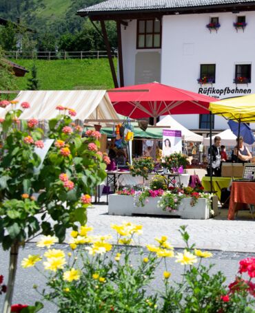 Tannbergermarkt im Freien mit bunten Blumen, Verkaufsständen und großen Sonnenschirmen und Im Vordergrund sind Blumen zu sehen.