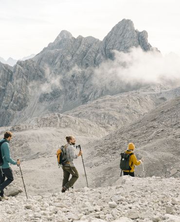 Drei Wanderer gehen auf einem felsigen Steig mit einem kargen Gebirgspanorama im Hintergrund. 