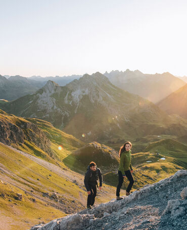 Eine Frau und ein Mann wandern bergauf während im Hintergrund die Sonne über die Berggipfel scheint
