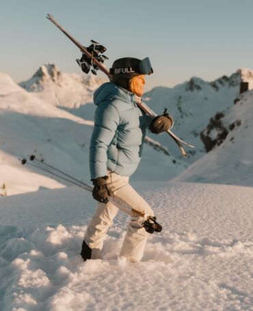eine Skifahrerin läuft durch den Tiefschnee mit ihren Ski auf den Schultern, bei Morgensonne