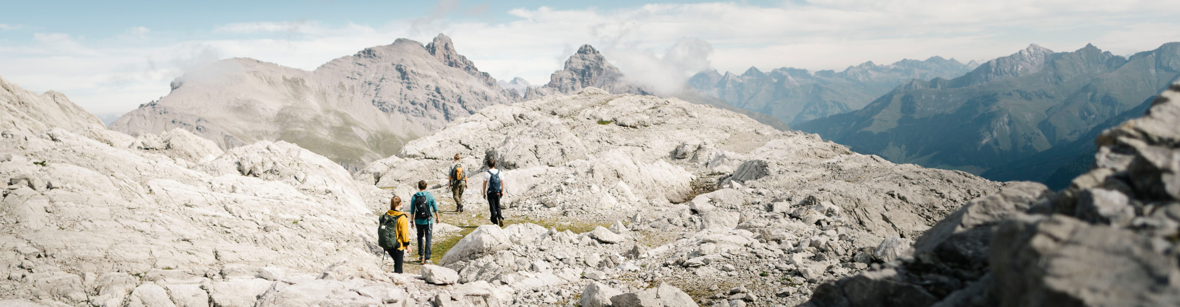 Vier Wanderer gehen entlang eines Steiges durch eine karge Felslandschaft. Im Hintergrund erstreckt sich ein weitläufiges Bergpanorama.