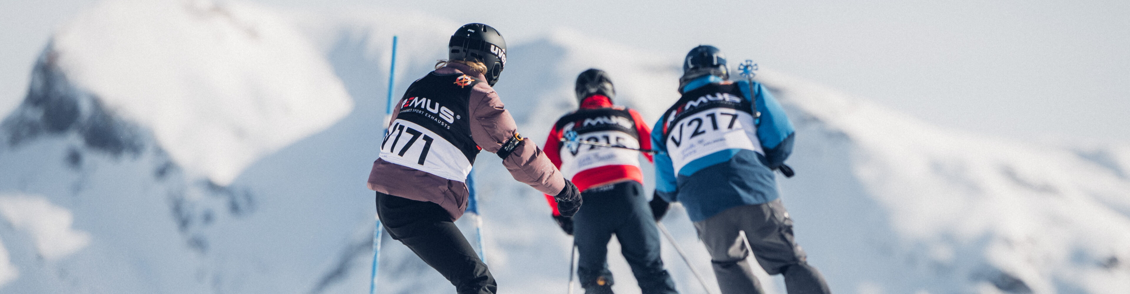 Zwei Skifahrer und ein Snowboarder fahren mit ihren Startnummern-Bibs bei strahlend blauem Himmel die verschneite Strecke des Weißen Rings hinunter