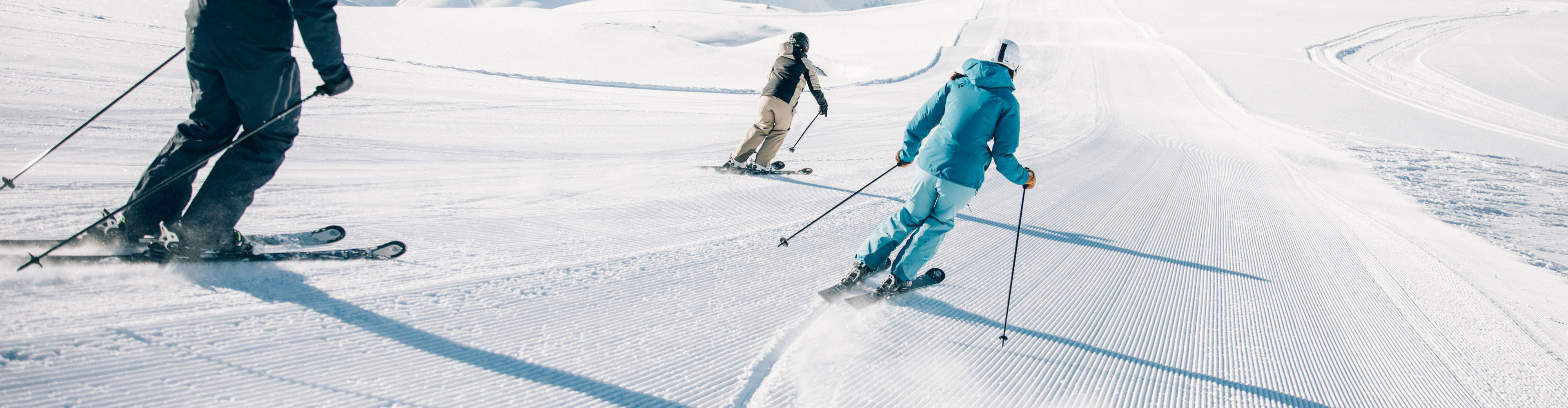 drei Skifahrer fahren bei klarem Sonneschein über die frisch präparierte Piste, mit traumhaften Bergkulissen im Hintergrund
