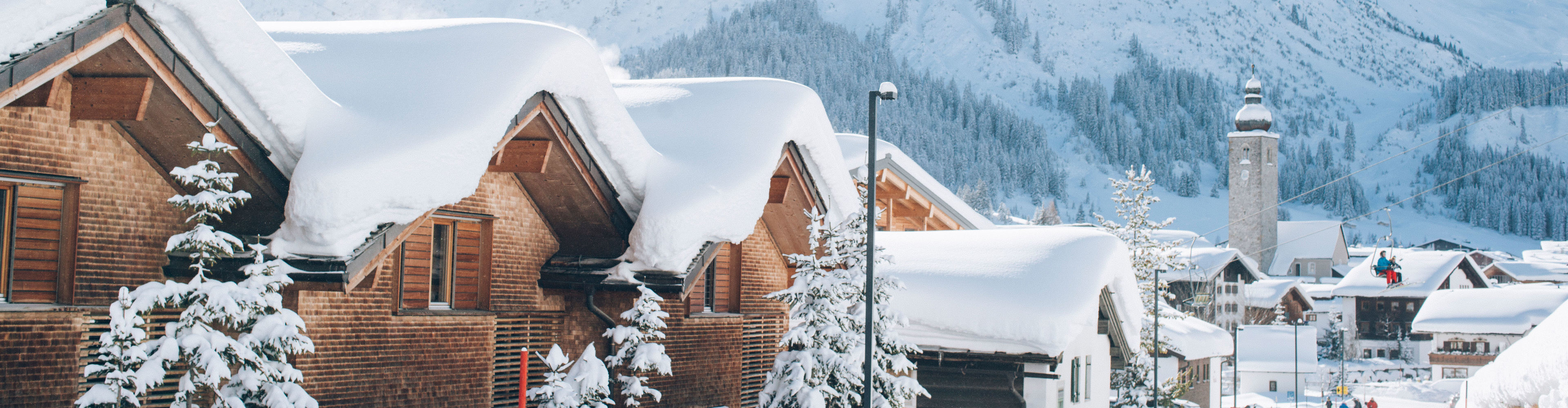 mehrere schneebedeckte Holzhäuser stehen vor der traumhaften Winterkulisse, im Hintergrund sieht man Lech und das Omeshorn