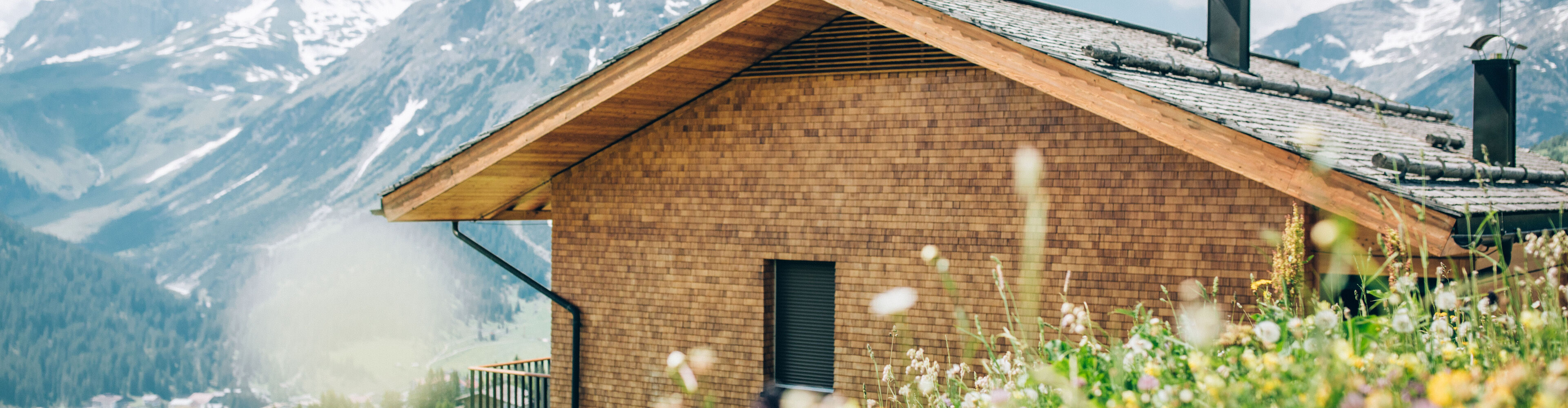 Bunte Frühlingswiese mit einem Holzhaus und einem steinigen Bergpanorama im Hintergrund