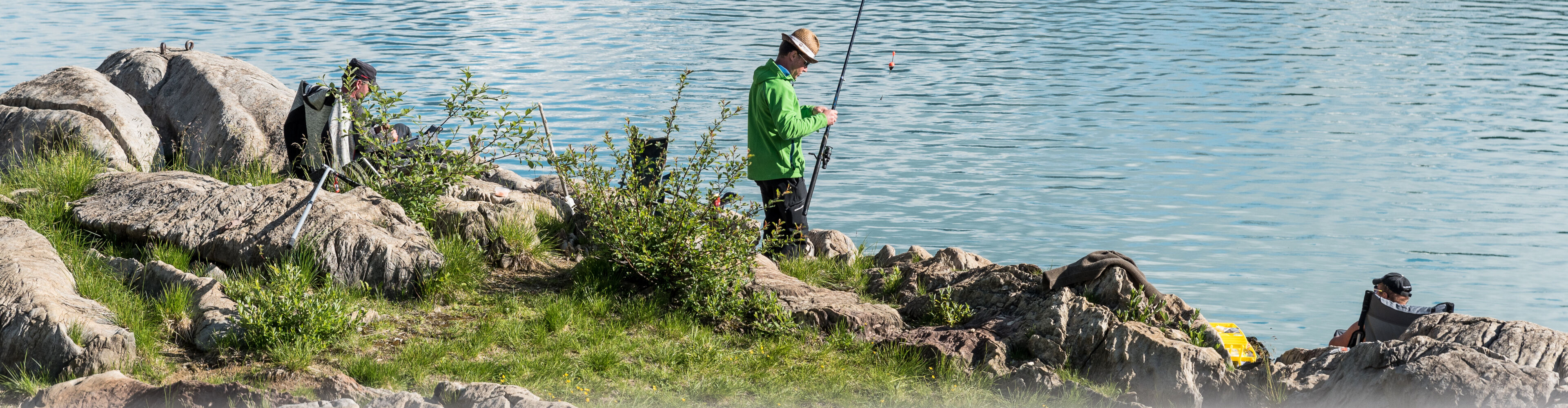 ein Fischer steht mit seiner Angel in der Hand auf der grünen Wiese vor dem blauen Fischersee