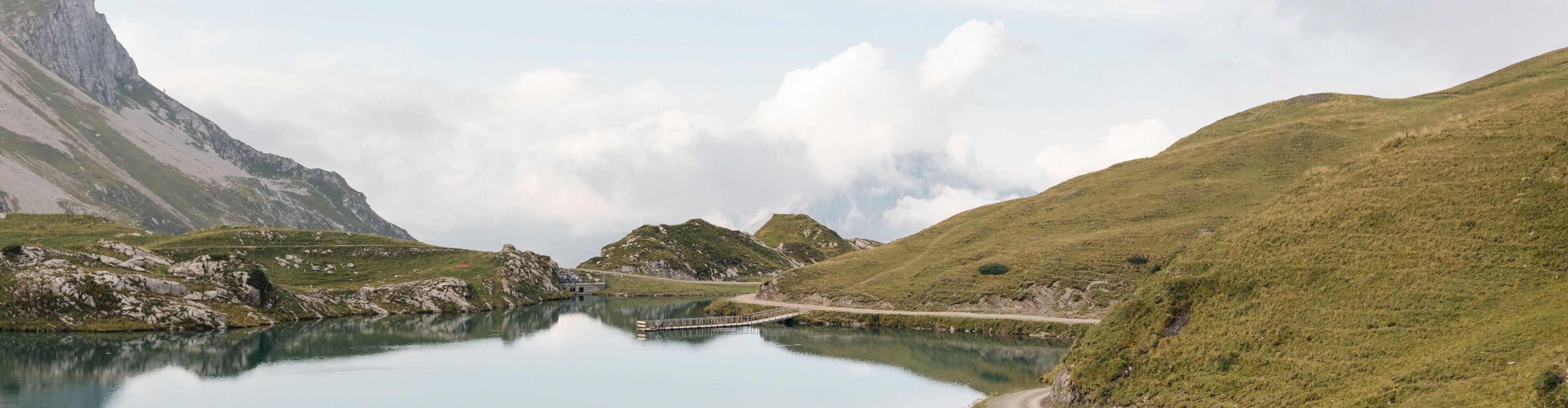 Ein Wanderweg verläuft entlang eines Gebirgssees. Im Hintergrund ist ein wolkenverhangenes Bergpanorama.