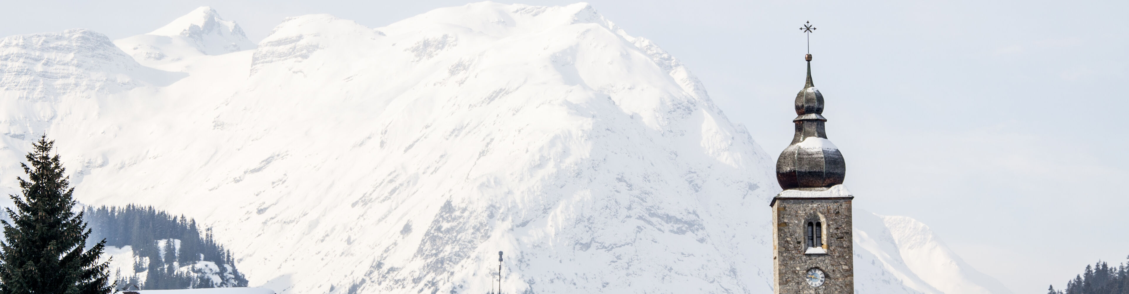 Verschneite Landschaft im Hintergrund und im Vordergrund die Kirche sowie zwei Häuser in Lech zu sehen