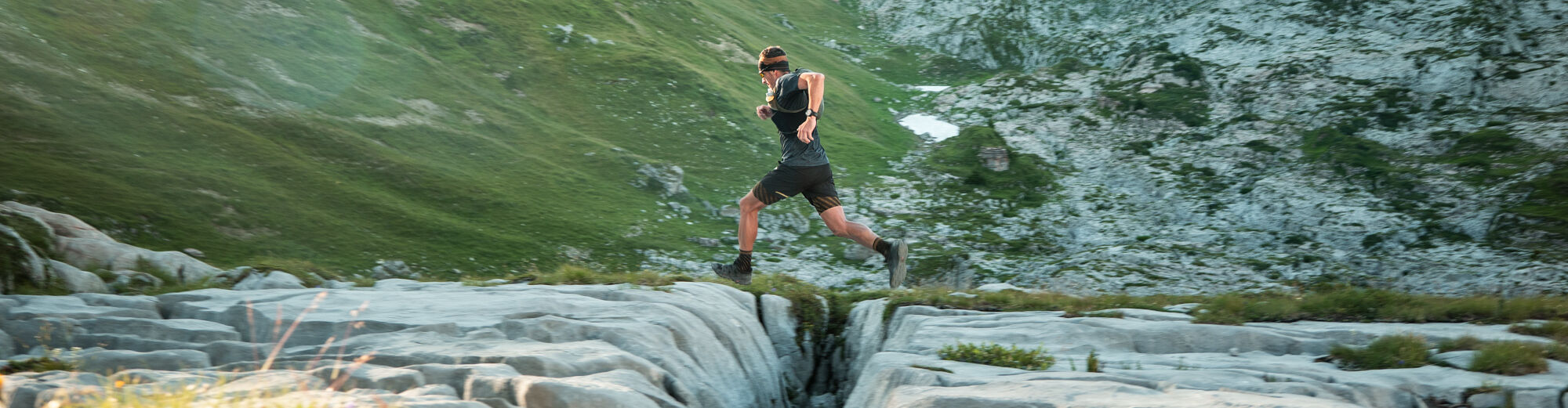 Trailrunner springt über eine tiefe Felsspalte im Karstgestein, im Hintergrund grüne Berghänge und zerklüftete Felsformationen im warmen Abendlicht.