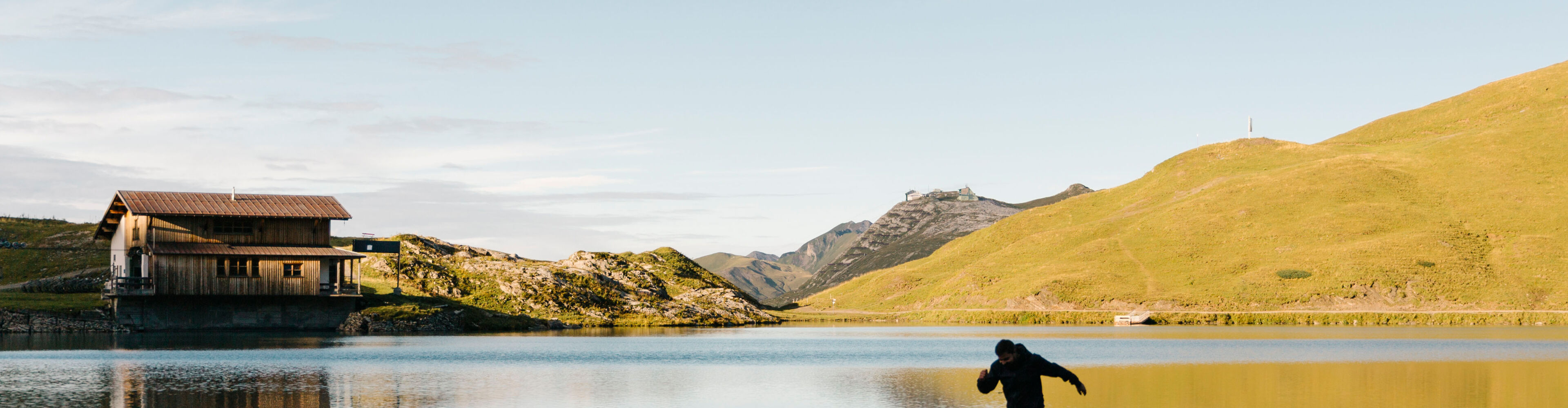 Ein Wanderer geht entlang eines Gebirgssees. Im Hintergrund wird eine Hütte sowie die umliegende Landschaft in das Licht der Morgensonne getaucht.