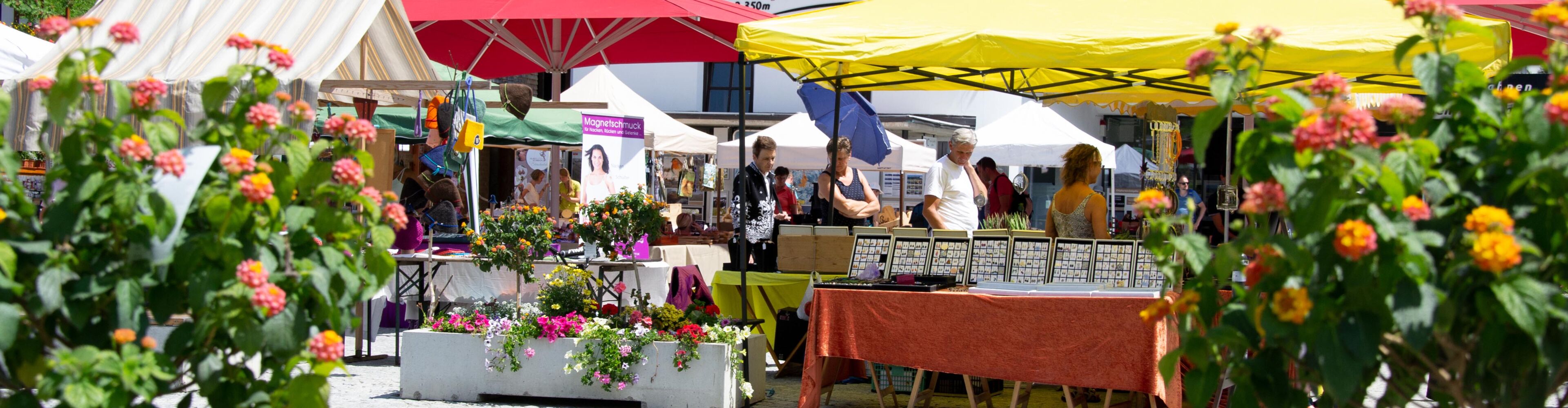 Tannbergermarkt im Freien mit bunten Blumen, Verkaufsständen und großen Sonnenschirmen und Im Vordergrund sind Blumen zu sehen.