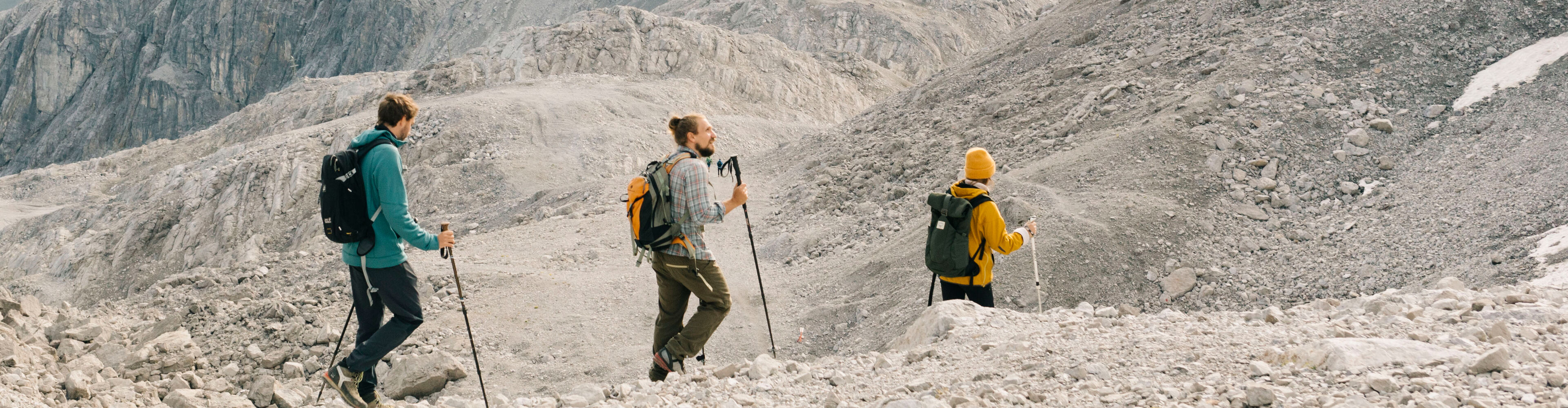 Drei Wanderer gehen auf einem felsigen Steig mit einem kargen Gebirgspanorama im Hintergrund.