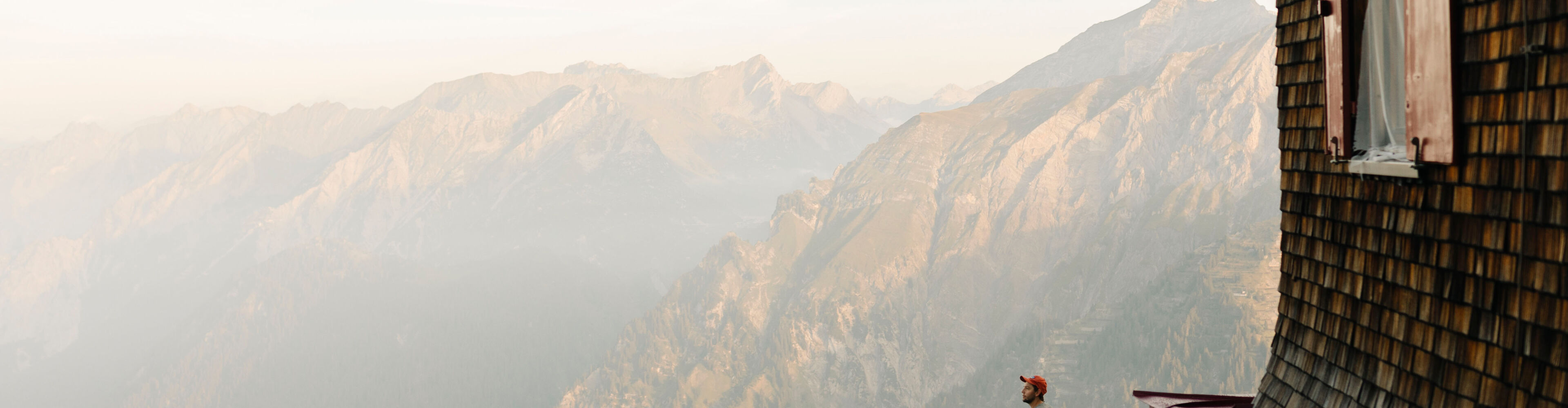 Ein Wanderer steht auf der Terrasse einer Hütte und blickt auf das Bergpanorama.