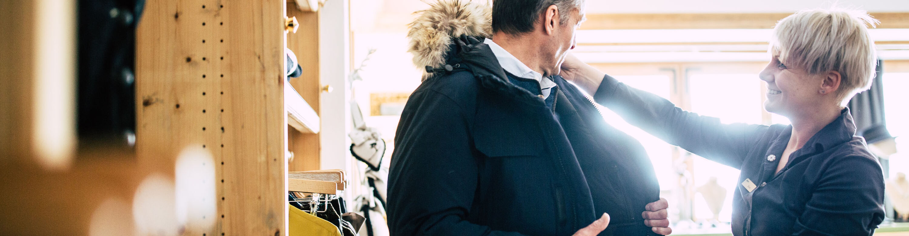 eine Verkäuferin berät einen Kunden im Geschäft, während er gerade eine Winterjacke anprobiert, im Hintergrund sieht man ausgestellte Kleidungsstücke des Geschäfts