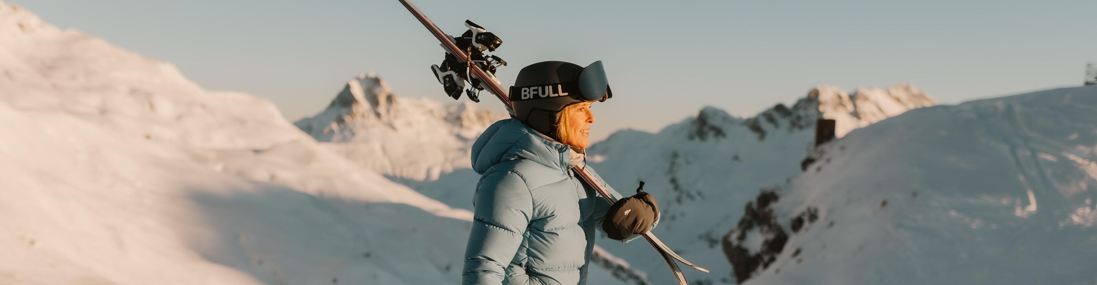 eine Skifahrerin läuft durch den Tiefschnee mit ihren Ski auf den Schultern, bei Morgensonne