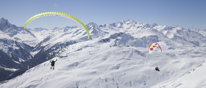 Zwei Paragleiter von hinten zu sehen und im Hintergrund eine große weite zu sehen mit dem Skigebiet und Berggipfeln