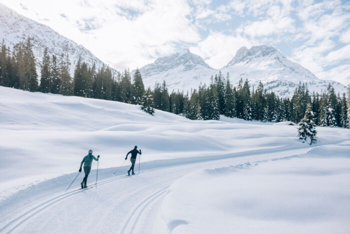 zwei Langläufer laufen über die Loipe im Zugertal während man im Hintergrund die verschneite Winterlandschaft sieht