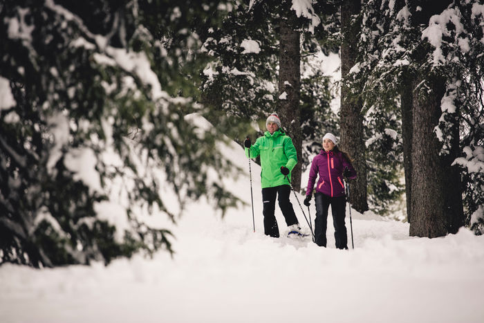 zwei Winterwanderer laufen durch die verschneite Winterlandschaft, im Hintergrund sieht man die schneebedeckten Nadelbäume